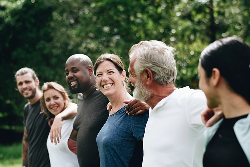 A diverse group of six adults stand close together outdoors, smiling and talking with their arms around one another.